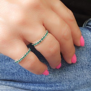 May include: Two silver rings with a row of small, green gemstones. The rings are worn on the fingers of a hand.