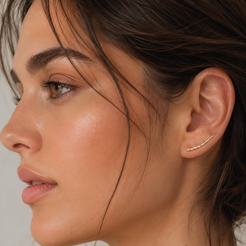 May include: A close-up of a person's face with a gold ear climber earring. The earring curves along the ear, featuring a textured surface. The skin tone is fair, and the hair is dark brown.