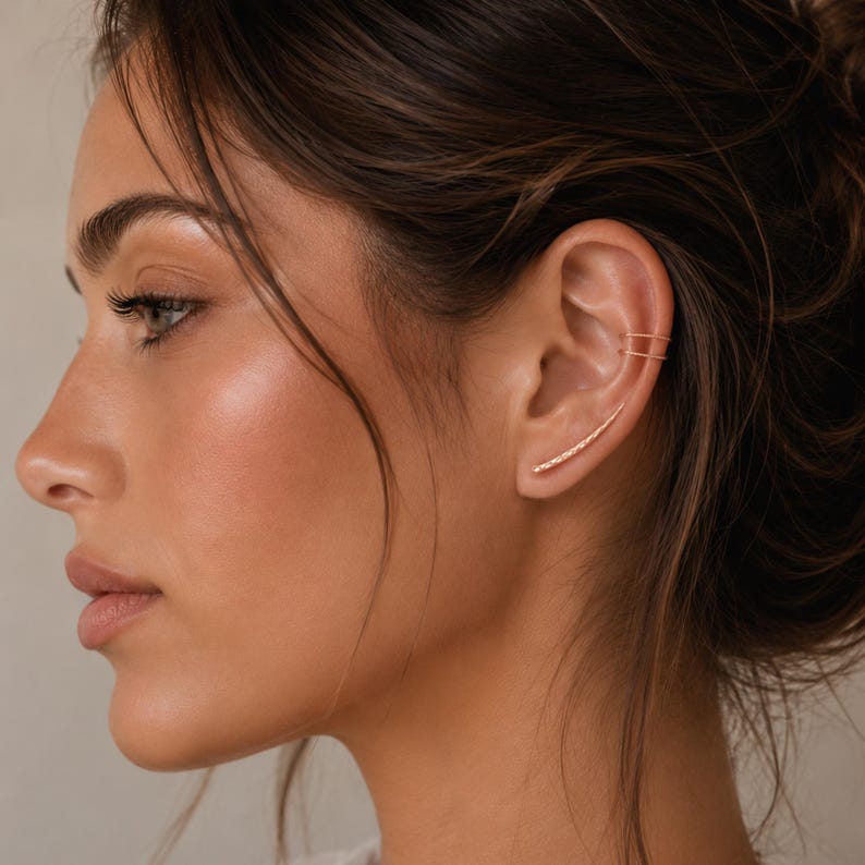 May include: A side profile of a person wearing gold-colored ear jewelry. The jewelry includes a curved ear cuff and a small stud earring. The ear cuff has a line of small, sparkling details. The person has dark hair and a fair complexion.