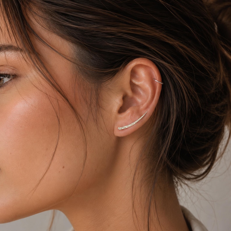 May include: A pair of silver ear climbers. The earrings curve upwards along the ear, featuring a textured surface. The design is minimalist and elegant, suitable for everyday wear.