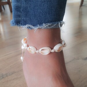 May include: A white shell anklet with a white string and a white bead. The anklet is on a person's ankle.