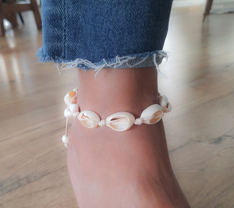 May include: A white shell anklet with a white string and a white bead. The anklet is on a person's ankle.