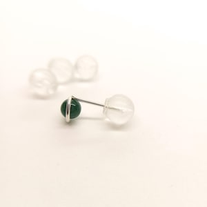 May include: A single stud earring with a small, round, dark green stone set in a silver metal band. The earring post is visible, along with a clear, round earring back. Additional earring backs are blurred in the background.