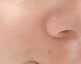 May include: Close-up of a person's nose with a small, silver-colored stud earring. The earring is a simple, heart-shaped design. The skin is fair-toned and smooth, with a few freckles visible. The focus is on the nose and the jewelry.