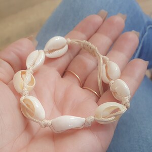 May include: A white shell bracelet with a beige cord. The bracelet is made of natural seashells and is tied with a simple knot.