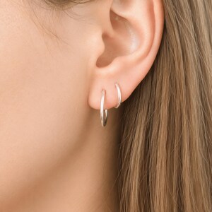 May include: Two small, polished silver hoop earrings worn in the earlobe. The earrings are simple, circular bands with a smooth, reflective metallic finish. One hoop is positioned slightly lower than the other, creating a layered appearance.