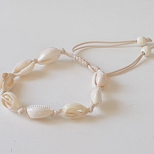 May include: A white seashell bracelet with a cream-colored cord. The bracelet is made of small, oval-shaped seashells strung together with a simple knot.