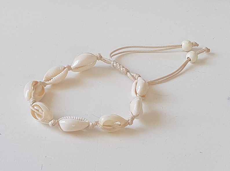 May include: A white seashell bracelet with a cream-colored cord. The bracelet is made of small, oval-shaped seashells strung together with a simple knot.