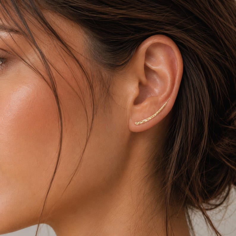 May include: A gold ear climber earring with a textured surface is shown on the ear. The earring curves upward along the ear's edge. The skin tone is light with dark brown hair.