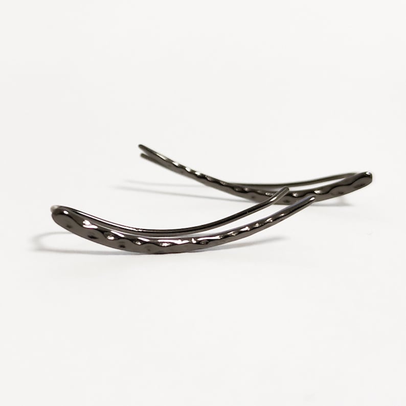 May include: A pair of dark gray ear climbers with a hammered texture. The earrings curve upwards, with a sleek, modern design. The jewelry is set against a white background.