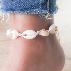 May include: A white seashell anklet with a beige cord. The anklet is tied around a person's ankle.