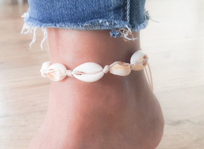 May include: A white seashell anklet with a beige cord. The anklet is tied around a person's ankle.