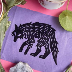 Peut inclure: Une estampe de loup noir sur fond violet. Le loup est représenté dans une pose stylisée en train de marcher. L'estampe est sur un papier texturé, entourée de plantes et d'un fond rose et blanc.