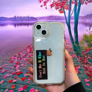 May include: Clear iPhone case with a black and iridescent sticker that reads "Don't Be A Thot Hell Is Hot". The phone case is held in front of a scenic background of a lake and fall foliage.