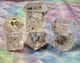 Puede incluir: Dados de resina transparente y facetada con naipes incrustados. Los dados presentan varios palos de cartas y figuras, creando un accesorio de juego único y decorativo. Los dados se muestran sobre una superficie colorida y estampada.