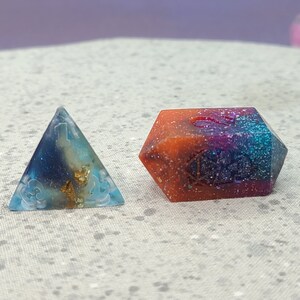 May include: Two handmade resin dice, one a pyramid and the other a multi-sided die. The pyramid is blue, gold, and white with a cloud design. The multi-sided die is orange, purple, and blue with glitter and a number 5.