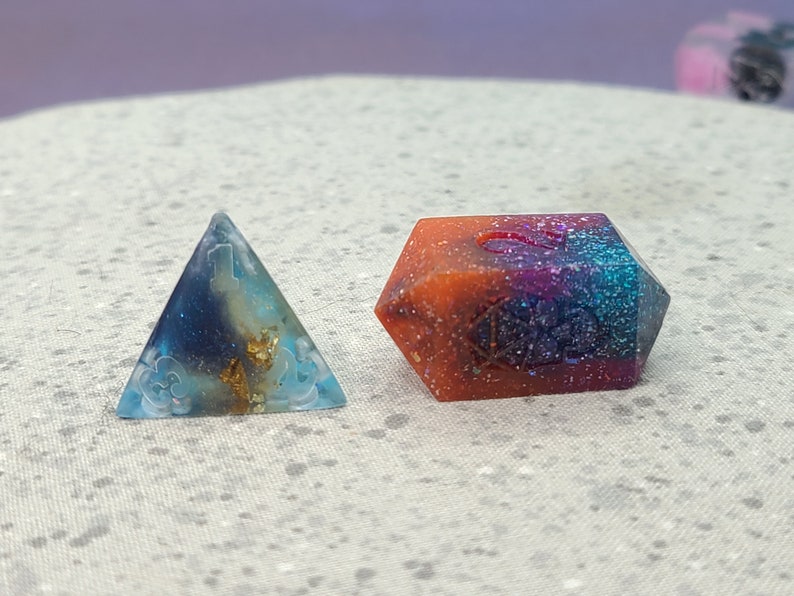 May include: Two handmade resin dice, one a pyramid and the other a multi-sided die. The pyramid is blue, gold, and white with a cloud design. The multi-sided die is orange, purple, and blue with glitter and a number 5.