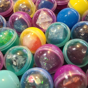 May include: A collection of colorful plastic capsules containing various dice with glitter and iridescent finishes. The capsules are arranged in a random pattern, showcasing the different colors and designs of the dice.