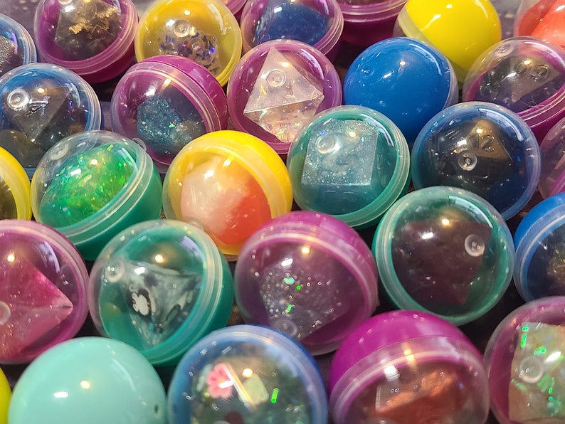 May include: A collection of colorful plastic capsules containing various dice with glitter and iridescent finishes. The capsules are arranged in a random pattern, showcasing the different colors and designs of the dice.