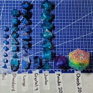 May include: A collection of blue and iridescent dice of various sizes, including mini, standard, large, jumbo, and mondo 20. The dice are arranged in rows on a blue grid background. There is also a large, multi-colored, iridescent dice with the text "Chonk 20" on it.