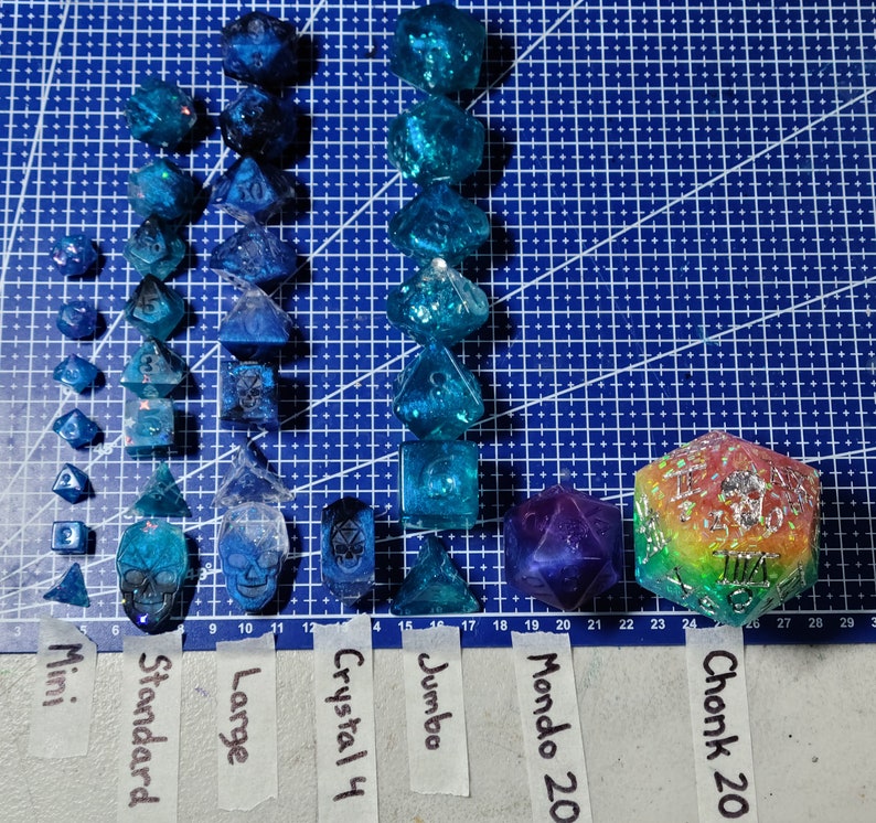May include: A collection of blue and iridescent dice of various sizes, including mini, standard, large, jumbo, and mondo 20. The dice are arranged in rows on a blue grid background. There is also a large, multi-colored, iridescent dice with the text "Chonk 20" on it.