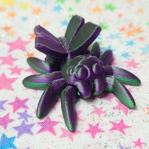 Puede incluir: Una araña impresa en 3D con alas iridiscentes de color púrpura y verde. La araña tiene una superficie detallada y texturizada y ojos grandes y redondos.