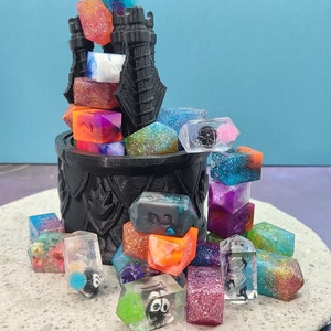 May include: A collection of colorful, resin dice in various shapes and sizes, spilling from a black, decorative cauldron-shaped container. The dice feature glitter, swirls, and embedded designs. The background is a light blue.