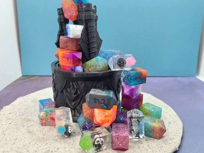 May include: A collection of colorful, resin dice in various shapes and sizes, spilling from a black, decorative cauldron-shaped container. The dice feature glitter, swirls, and embedded designs. The background is a light blue.