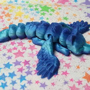 May include: A blue and teal 3D printed dragon figurine with wings, lying on a white background with a rainbow star pattern.