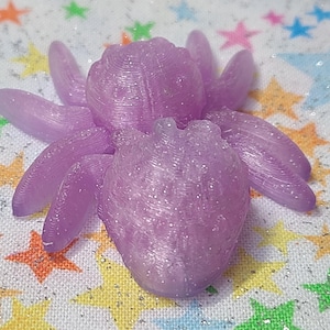 Puede incluir: Una figurita de araña de color morado y brillante con una superficie texturizada detallada. La araña está hecha de plástico y tiene ocho patas.