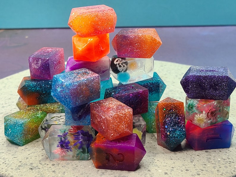 May include: A collection of colorful, glitter-infused resin dice. The dice are various colors, including orange, purple, blue, and green, with embedded designs and glitter. Some dice have floral inclusions, and others have visible numbers.