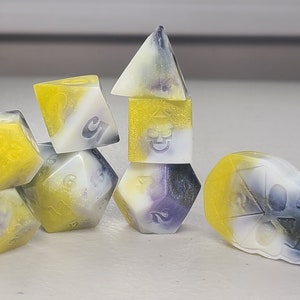 May include: A set of seven dice with a yellow, white, and purple swirl pattern. The dice are shaped like a cube, a pyramid, and a skull. The skull die has a black skull design on one side.