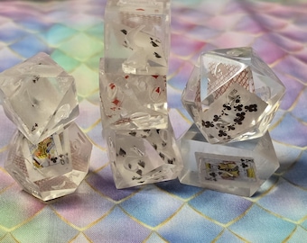 Puede incluir: Dados geométricos transparentes con diseños de naipes incrustados. Los dados están dispuestos sobre una superficie colorida y estampada. Las cartas presentan palos como tréboles, diamantes y picas.