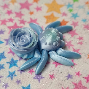 Puede incluir: Una araña impresa en 3D de color azul claro con una rosa azul claro en la espalda. La araña y la rosa están sobre un fondo blanco con un patrón de estrellas de colores.