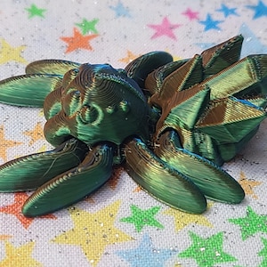 Puede incluir: Una criatura articulada con forma de araña, impresa en 3D, de color verde e iridiscente, con una espalda espinosa.