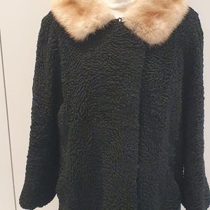 May include: A black, textured coat with a light brown fur collar. The coat is displayed on a white mannequin. The coat has long sleeves and a button closure. The fur collar adds a touch of luxury to the classic design.