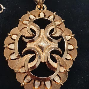 May include: Gold-toned pendant necklace with a large, ornate circular pendant. The pendant features a central starburst design and curved, petal-like elements. The chain is a silver-colored metal.