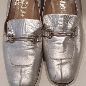 May include: A pair of silver-colored leather loafers with a metallic finish. The shoes have a square toe design and a silver buckle detail across the top. The interior shows a gold logo that reads "Miotto" and "made in Italy".