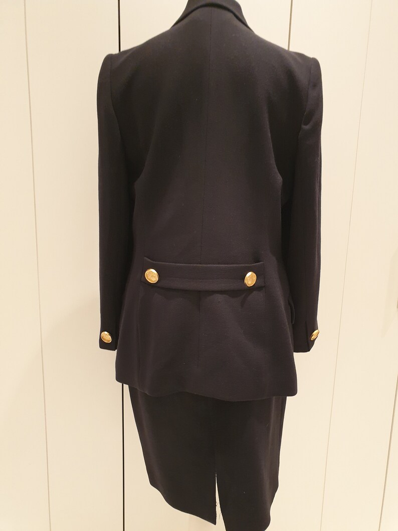 May include: A black blazer and skirt suit with gold buttons. The blazer has long sleeves and a double-breasted closure. The skirt is knee-length and has a straight silhouette.