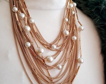 May include: A multi-strand gold-tone necklace with faux pearls. The necklace features multiple layers of delicate chains and bar accents, interspersed with large, round, white faux pearls. The necklace is displayed on a white surface, with a green, feathery texture in the background.