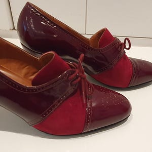 Può includere: Un paio di scarpe bordeaux da donna con un design bicolore. Le scarpe presentano una tomaia in vernice lucida e un pannello in camoscio sulla punta e sul collo del piede. Hanno una chiusura con lacci e un piccolo tacco.