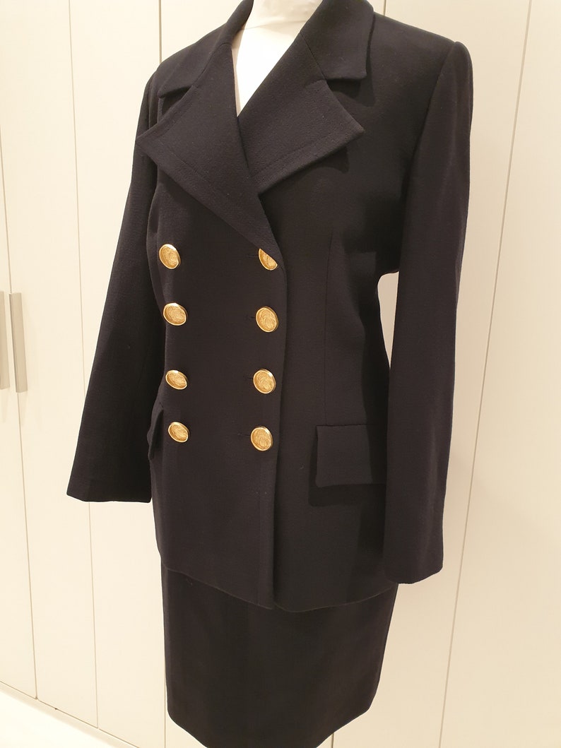 May include: A black double-breasted blazer with gold buttons and a matching skirt. The blazer has a classic collar and long sleeves. The skirt is knee-length and has a straight silhouette.