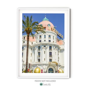 Könnte beinhalten: Ein weißes Gebäude mit einer rosafarbenen Kuppel und einem goldenen Schild mit der Aufschrift "Le Negresco" vor blauem Himmel. Im Vordergrund steht eine Palme.