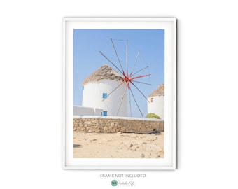 May include: Two white windmills with red blades stand against a blue sky. The windmills are topped with straw roofs and have blue accents. The windmills are surrounded by a stone wall and sand.