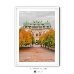 May include: A white building with a green roof stands in the middle of a path lined with trees. The trees have orange and yellow leaves. The building has a symmetrical design with multiple windows and a central entrance.