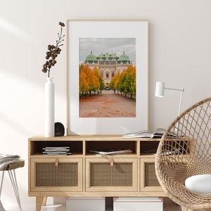 May include: A framed photograph of a building with a green roof and yellow walls. The building is surrounded by trees with orange and yellow leaves. The trees are in a row and lead to the building. The photo is hanging on a wall above a wooden cabinet with three drawers.
