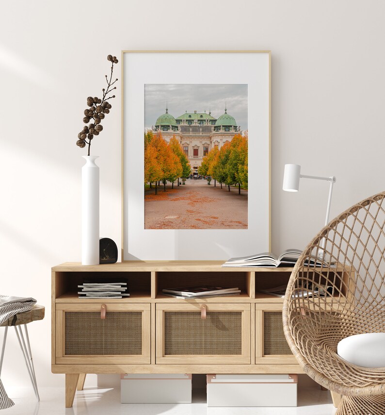 May include: A framed photograph of a building with a green roof and yellow walls. The building is surrounded by trees with orange and yellow leaves. The trees are in a row and lead to the building. The photo is hanging on a wall above a wooden cabinet with three drawers.