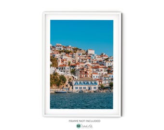 May include: A colorful cityscape with white buildings and red tile roofs on a hillside overlooking a blue body of water. The buildings are clustered together and have a variety of architectural styles. The water is calm and reflects the blue sky.