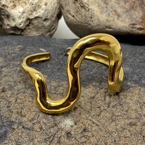 May include: A gold-toned ring featuring a continuous, fluid, wavy, or serpentine design. The metal surface is smooth and reflective, with an open-ended band structure that allows for adjustment. The ring has a polished finish and a curved, organic shape.