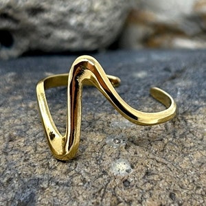 Peut inclure: Une bague en or avec un design ondulé abstrait. La bague est posée sur une roche grise.